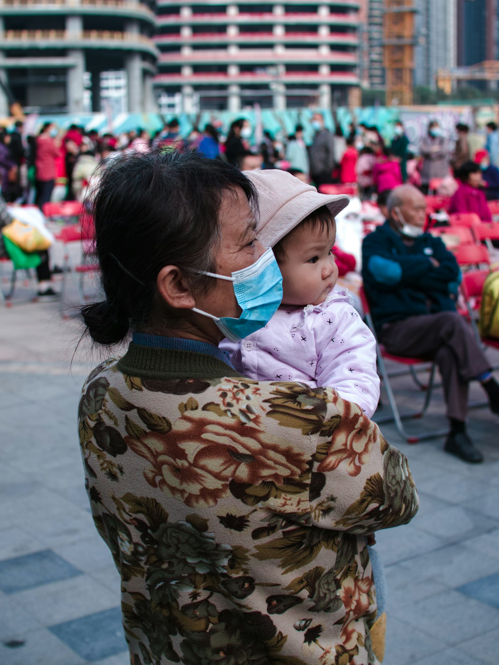 A grandmother wearing a face mask holding a baby outdoors in a public plaza