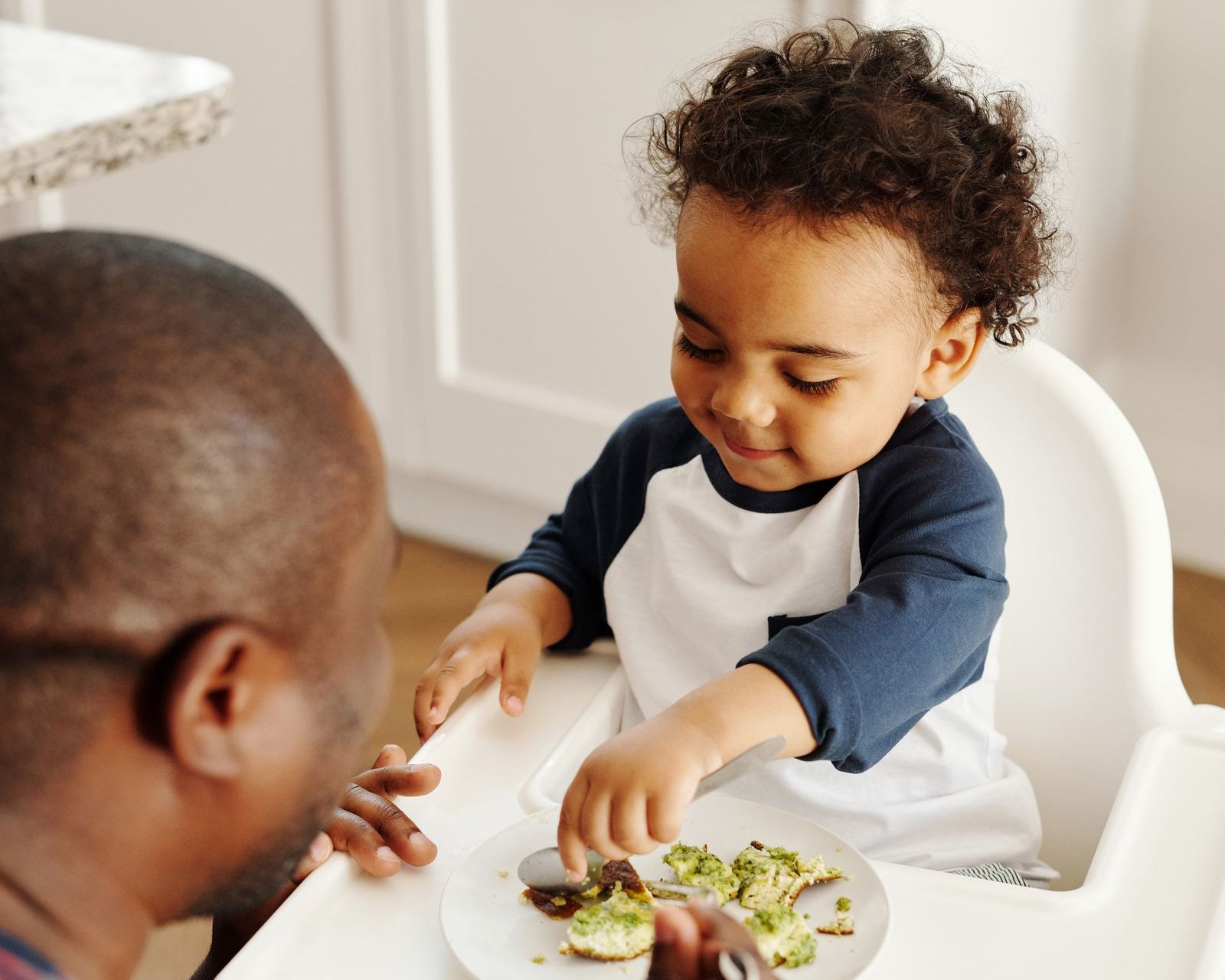 A toddler in a white high chair reaches for soft mashed avocado pieces on a plate while a parent sits close by