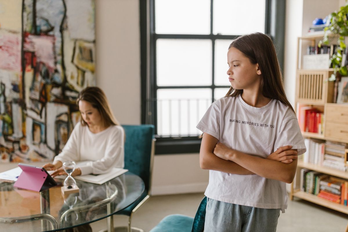 An upset child with arms crossed near a parent using a device