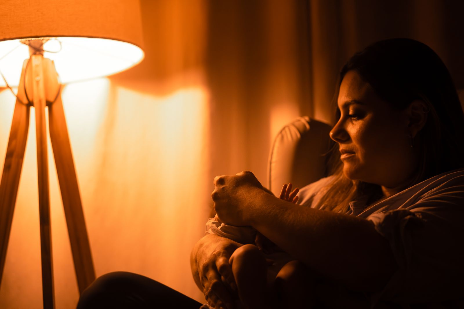 A mother sits in a chair beside a glowing lamp in a dim room, gently cradling her baby during a night feed