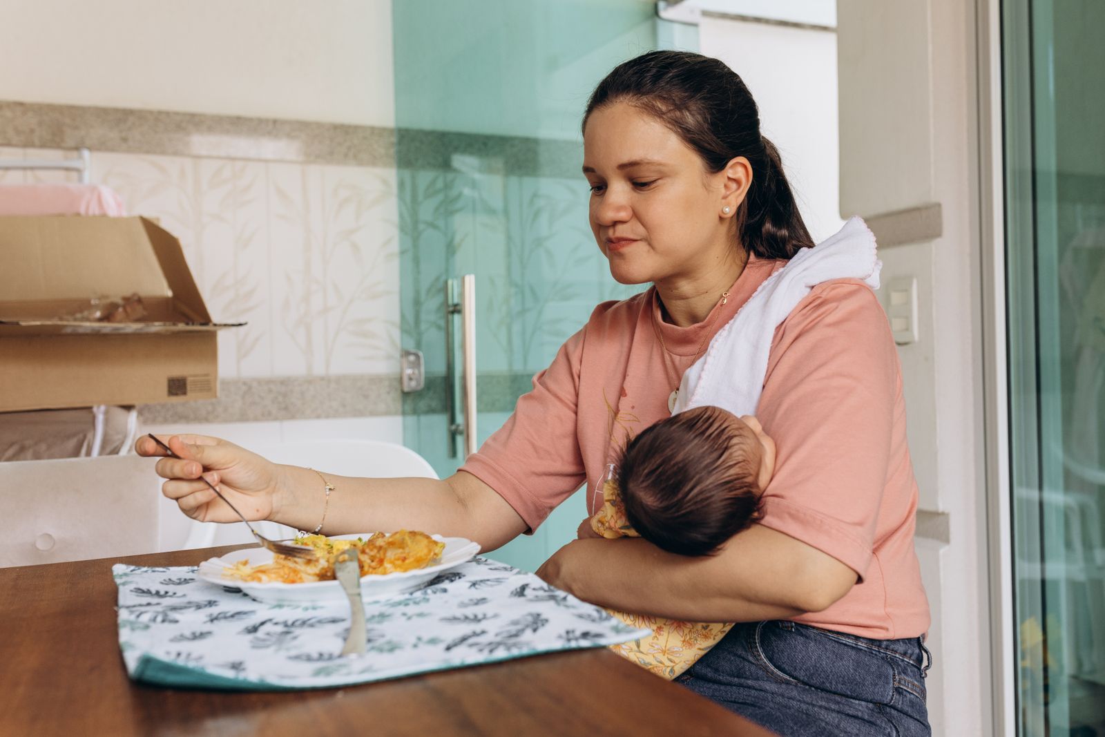 A mother sitting at a wooden table eating a plate of food with a fork while holding her newborn against her shoulder