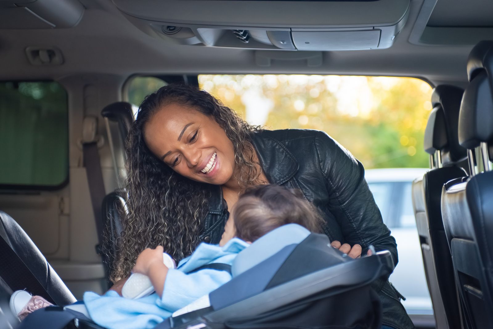 A mother leans in and smiles at her baby, who is settled in an infant car seat in the back seat of a van