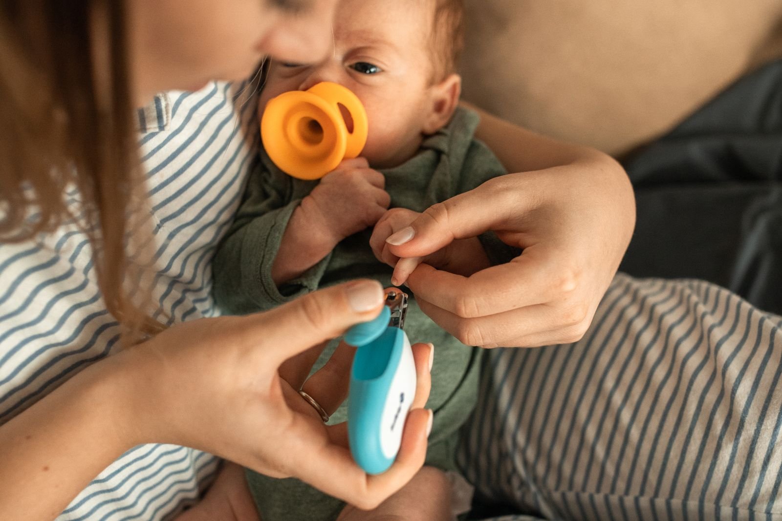 A mother holds her newborn on her lap and gently trims one fingernail with baby nail clippers while supporting the baby's hand