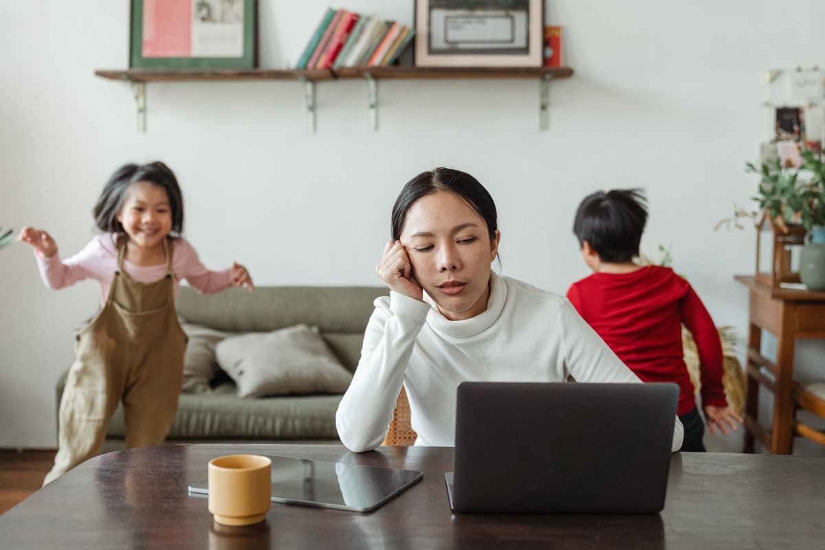 A mother resting her head in her hand while working at a laptop as her two children play behind her