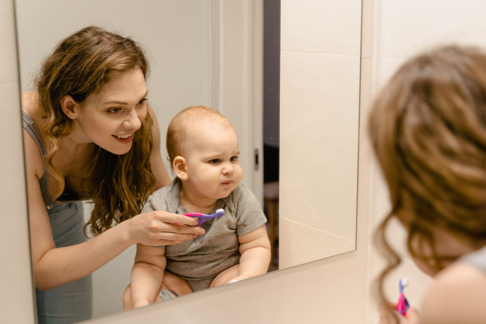 A parent helping a toddler brush their teeth in front of the bathroom mirror