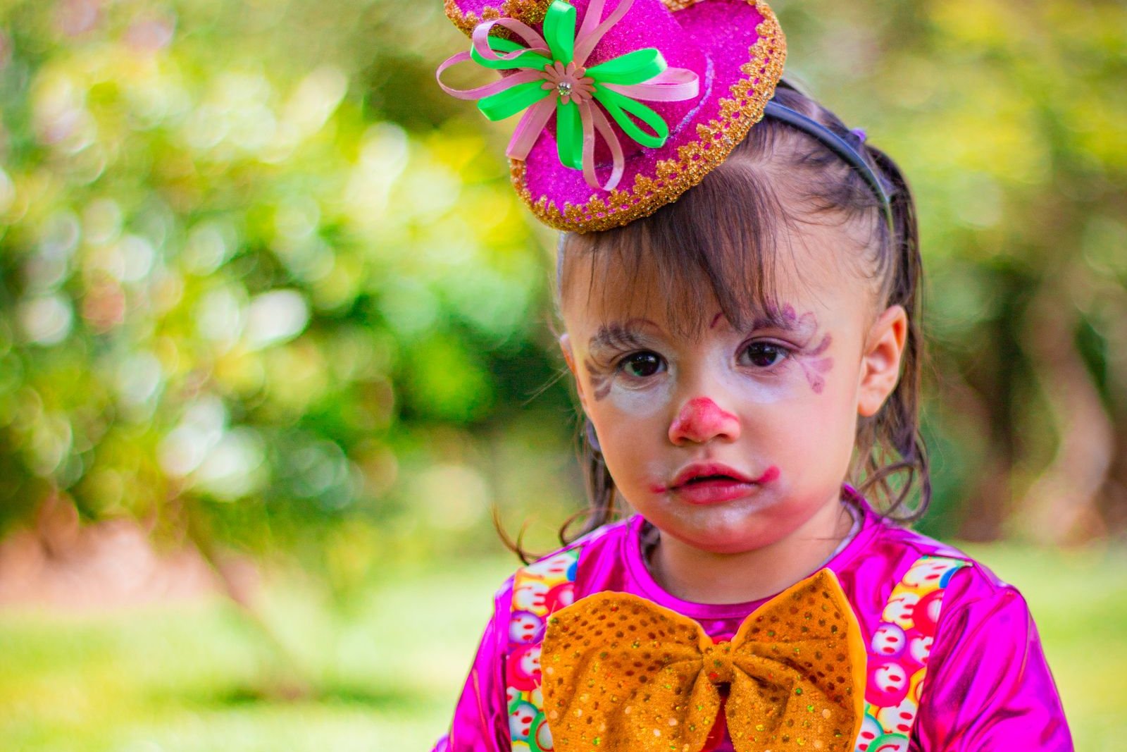 Toddler in a pink clown costume with a small glittery hat and light face paint, looking at the camera in a sunny garden
