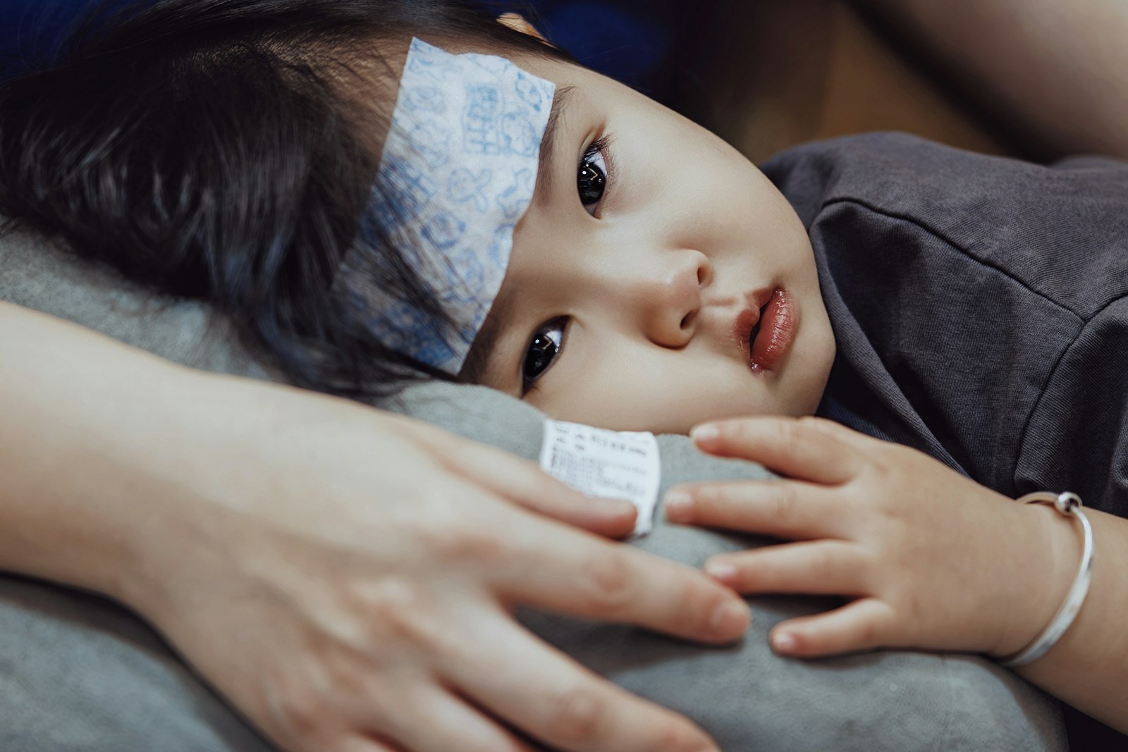 A sleepy toddler with a cooling patch on her forehead rests on a parent's lap while the parent's hand holds her close