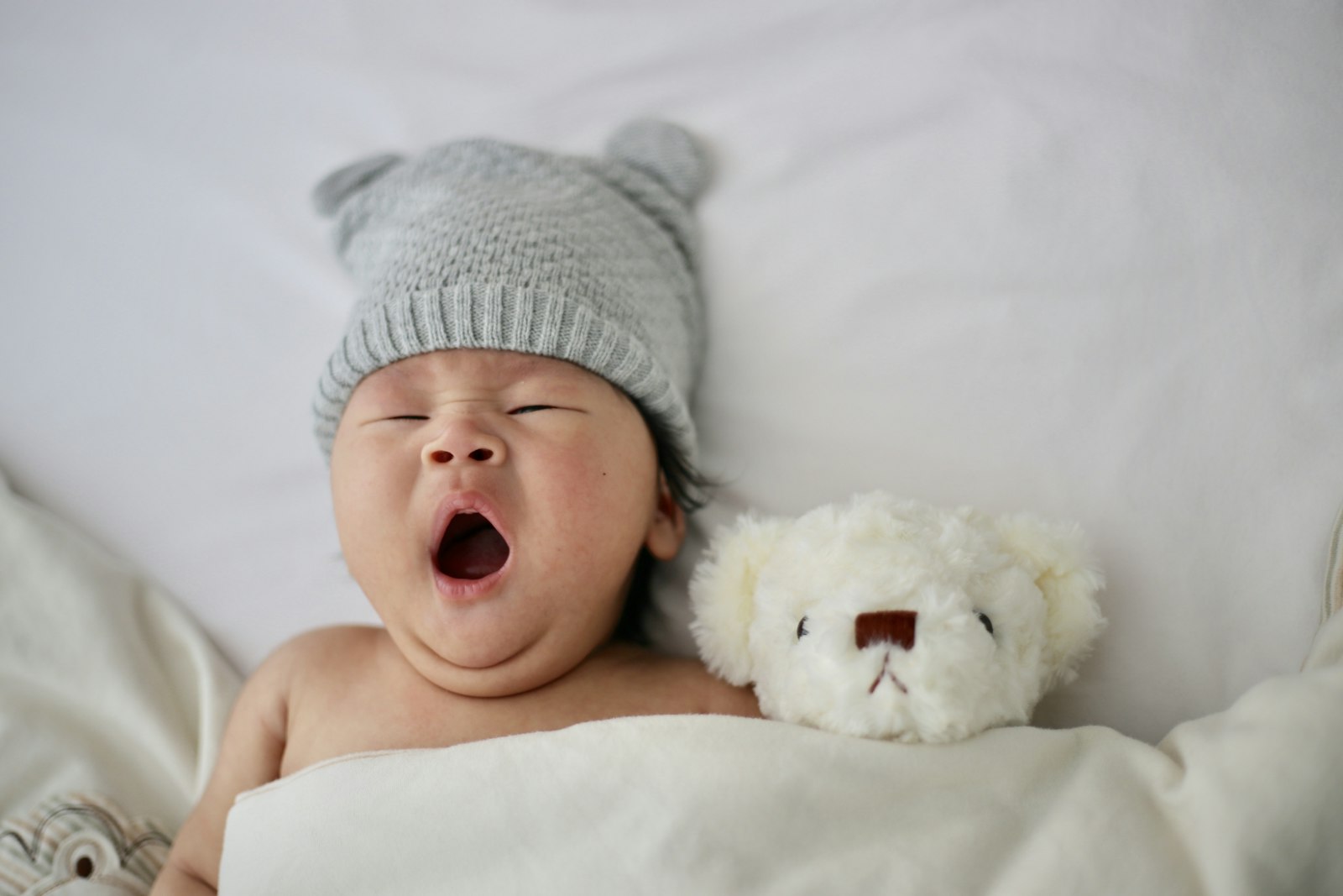 A yawning baby wearing a grey knit hat beside a white teddy bear